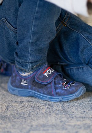 Blaue Stoffschuhe mit Klettverschluss, die den Text "POLICE" und eine Autografik zeigen, mit strukturiertem Design und blauer Sohle, kombiniert mit Jeans.
