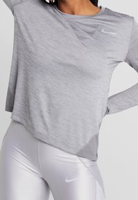 Person som bär en ljusgrå långärmad sportskofta och glänsande ljusgrå tights med Nike-logotyper på bröstet och låret.