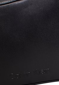 Czarna skórzana torba o gładkiej fakturze z wytłoczonym logo "Calvin Klein". Krawędzie zdobi kontrastowe przeszycie na górze.