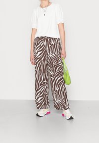 Femme portant un chemisier blanc à manches bouffantes, un pantalon large à imprimé zèbre marron et blanc, des baskets blanches avec des accents roses, et tenant un petit sac à main vert.