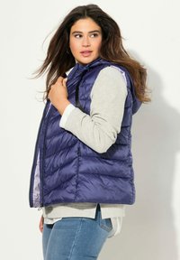 Izolowana kamizelka puchowa w ciemnofioletowym kolorze z zamkiem z przodu, kapturem i kieszeniami bocznymi. Ma wzór w zygzaki i lekką, błyszczącą fakturę.