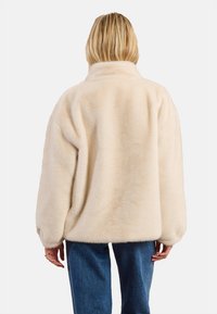 Giacca in pile color crema con colletto alto, vestibilità oversize e maniche lunghe. Tessuto morbido con cuciture minime, indossata sopra jeans blu.