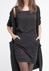 Schwarzes ärmelloses Oberteil kombiniert mit einem schwarzen Rock mit roten floralen Ausschnittdesigns, dazu ein langer schwarzer Cardigan und transparente Strumpfhosen.