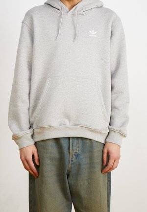 Osoba w jasnoszarej bluzie Adidas z kieszenią na przodzie i sznurkami, w połączeniu z wyblakłymi niebieskimi dżinsami, stoi na tle jednolitego tła.