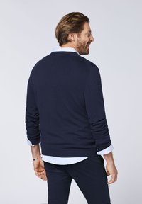 Navy gestrickter Pullover mit rundem Ausschnitt und gerippten Bündchen, getragen über einem hellblauen Hemd mit Kragen, das ein lässiges Rückenprofil zeigt.