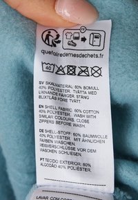 Verzorgingslabel op lichtblauwe stof, met wasinstructies in verschillende talen, samengesteld uit 60% katoen en 40% polyester.
