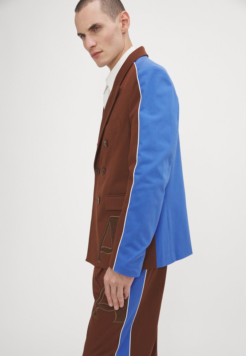 Tailleur marrone e blu con giacca doppiopetto, maniche blu a contrasto e dettagli dorati sui pantaloni. Texture liscia.