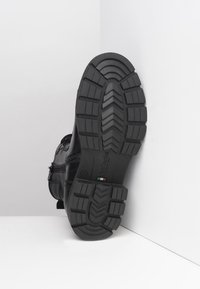 Černá kožená bota s robustní gumovou podrážkou, která má texturovaný vzor na podrážce a boční zip. Ozdobena kovovou sponou.