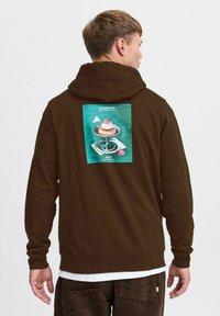 Brun hættebluse med stort grafisk print på ryggen, der viser en dessert på en glasstand, sat mod en teal baggrund. Blød tekstur, afslappet pasform.