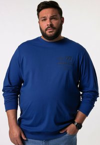 Niebieski długi rękaw dresowy wykonany z bawełny, z okrągłym dekoltem, ściągaczami na rękawach i nadrukiem z przodu. Noszony z niebieskimi dżinsami i zegarkiem.