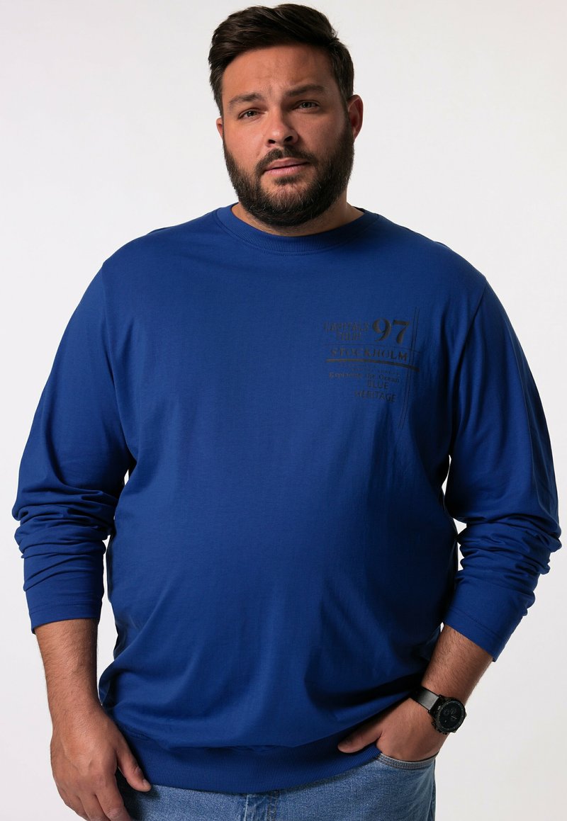 Niebieski długi rękaw dresowy wykonany z bawełny, z okrągłym dekoltem, ściągaczami na rękawach i nadrukiem z przodu. Noszony z niebieskimi dżinsami i zegarkiem.