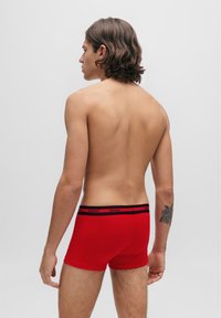 Boxer shorts rouges en coton avec une ceinture noire portant le nom de la marque "HUGO" en texte répétitif. Le modèle est vu de dos.