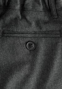 Tmavohnedá látka s textúrovaným povrchom, s malým čiernym gombíkom označeným "Clarks" a zadnou vreckom s lemovaním.