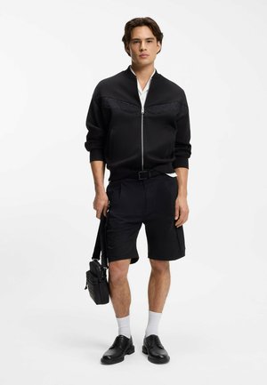 Jonge man met een zwarte jas met ritssluiting, zwarte korte broek, witte sokken, zwarte schoenen en een kleine zwarte schoudertas in zijn hand.