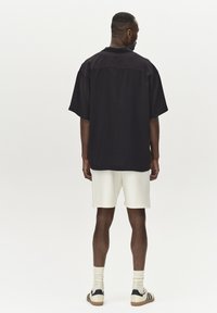 Svart kortärmad skjorta med avslappnad passform, i bomullstyg, med sidsprund. Matchad med vita shorts och krämfärgade strumpor, ståendes skor med ränder.