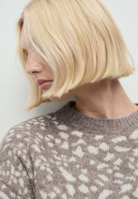 Pull léger beige avec un motif texturé en blanc, doté d'un col ras du cou et d'un matériau doux semblable à de la laine. Cheveux blonds courts et droits.