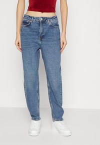 Jeans de denim azul claro com perna reta, cinco bolsos e acessórios em cobre. Usados com ténis brancos e um top vermelho curto.