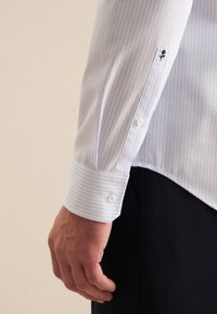 Camicia da uomo a righe blu chiaro con polsino bianco e chiusura a bottone, con un ricamo di una rosa nera sull manica e una texture di tessuto morbida.