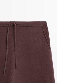 Pantalons de jogging bordeaux avec une taille à cordon de serrage, des poches latérales et une texture douce et lisse. Design simple sans motifs ni accents.