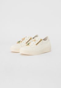 Las zapatillas de lona blancas presentan una suela acanalada, detalles en dorado en los cierres y elementos decorativos como adornos verdes y dorados.