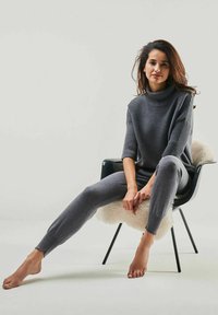 Grauer Strickrollkragenpullover mit gerippter Textur, kombiniert mit passenden enganliegenden Leggings. Model sitzt barfuß in einem schwarzen Stuhl.