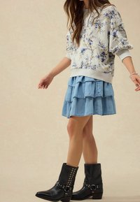 Sweat-shirt gris à imprimé floral avec manches bouffantes, jupe en denim bleu clair superposée, et bottines noires cloutées.