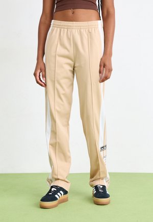 Person iført beige Adidas træningsbukser med hvide striber og sorte Adidas sneakers med gummisåler, stående på grøn gulv.