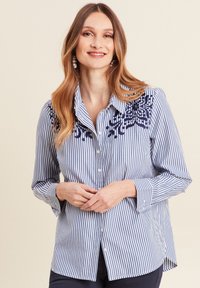 Chemise boutonnée en coton avec des rayures verticales bleu et blanc, ornée d'une broderie marine complexe sur l'épaule gauche et à manches longues.