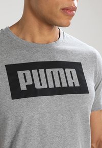 Harmaa puuvillapaita, jossa on suuri musta suorakulmainen logo, jossa on teksti "PUMA" rohkeilla kirjaimilla rinnassa. Lyhyet hihat, pyöreä kaula-aukko.