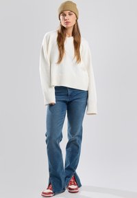 Pull court crème clair avec des poignets côtelés, associé à un jean bleu évasé avec fentes latérales et des baskets rouges avec des accents blancs.