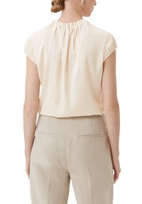 Satin blouse in light beige with short, pleated sleeves and gathered neckline. Paired with high-waisted, light beige trousers featuring back pockets.