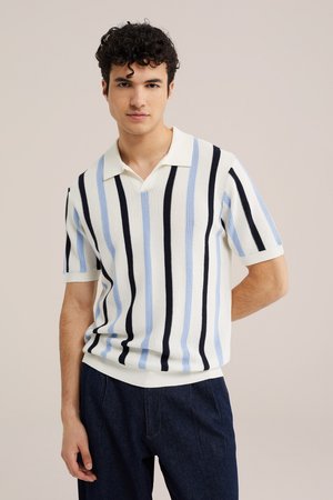 Jonge man met krullend haar die een witte gebreide polo met korte mouwen draagt met verticale blauwe en zwarte strepen en een donkere broek.