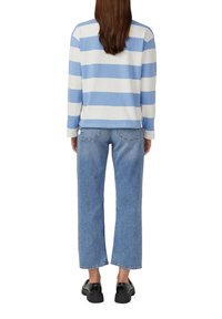 Person von hinten gesehen, der ein langärmliges Hemd mit breiten blauen und weißen Streifen, hellblaue gerade geschnittene Jeans und schwarze Schuhe trägt.