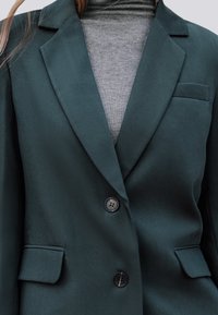Donkergroene blazer van gladde stof, met een notch-kraag, twee voorzakken en twee knopen. Draagt over een grijze coltrui.