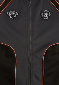 Zwarte zip-up jas met oranje piping, voorzien van een gestileerd apengezicht logo aan de linkerkant en een ronde BB-embleem op de rechterborst.