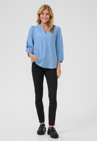 Lichtblauwe blouse met lange mouwen, een V-hals, geplooide manchetten en een relaxte fit, gecombineerd met zwarte skinny jeans en zwarte plateau schoenen.