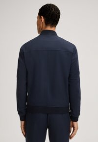 Navy blauwe bomberjack met een gladde textuur, ribbelstof bij de mouwen en taille. Beschikt over een kleine kraag en verticale naaddetails op de rug.