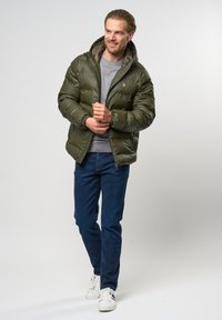 Aliejinės žalios spalvos pūkinė striukė su kapišonu, su užtrauktuko uždarymu, juostelėmis ant riešų ir minkšta pamušalu. Dėvima ant pilkos megztinio ir mėlynų džinsų.