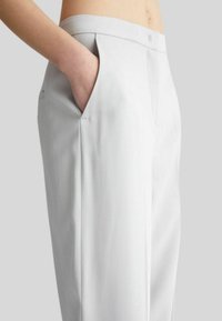 Pantaloni grigi chiari su misura con una texture liscia, dotati di una tasca laterale e una vestibilità strutturata. Cuciture visibili e linee pulite.