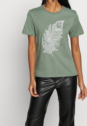 Osoba ubrana w zielony t-shirt z dużym, białym, szczegółowym wzorem liścia, w połączeniu z błyszczącymi czarnymi skórzanymi spodniami, stojąca na jednolitym tle.
