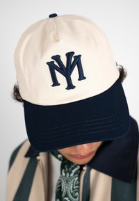 Casquette de baseball beige et noire avec logo "NY" superposé portée par une personne regardant vers le bas, visage et vêtements partiellement visibles.