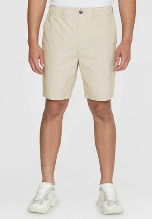 Mænd iført beige korte knælange shorts med knap og elastisk talje, kombineret med hvide sneakers og en hvid skjorte.