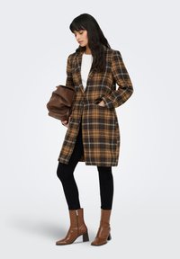 Palton în carouri maro și negru cu un design croitorit, având revere decupate și buzunare laterale. Asortat cu pantaloni negri și cizme maro cu toc.