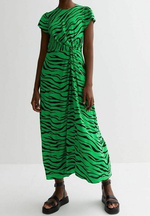 Woman wearing a green and black zebra-striped midi dress with cap sleeves and black platform sandals, standing against a plain white background.