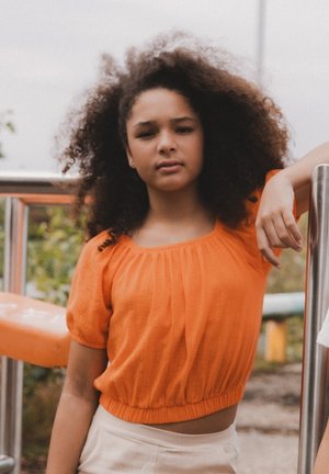Oranje top met korte mouwen, met een geribbelde textuur en een gezamelde onderkant, gecombineerd met lichte broek. De achtergrond bevat groenblijvende planten en een metalen leuning.