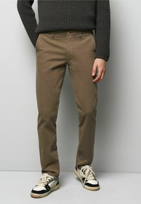 Bruine slim-fit broek van katoen met een gladde textuur, voorzien van zijzakken en een enkele knoopsluiting, gedragen met zwart-witte sneakers.