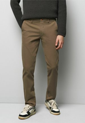 Braune Slim-Fit-Hosen aus Baumwolle mit glatter Textur, ausgestattet mit Fronttaschen und einem Knopfverschluss, kombiniert mit schwarz-weißen Sneakers.