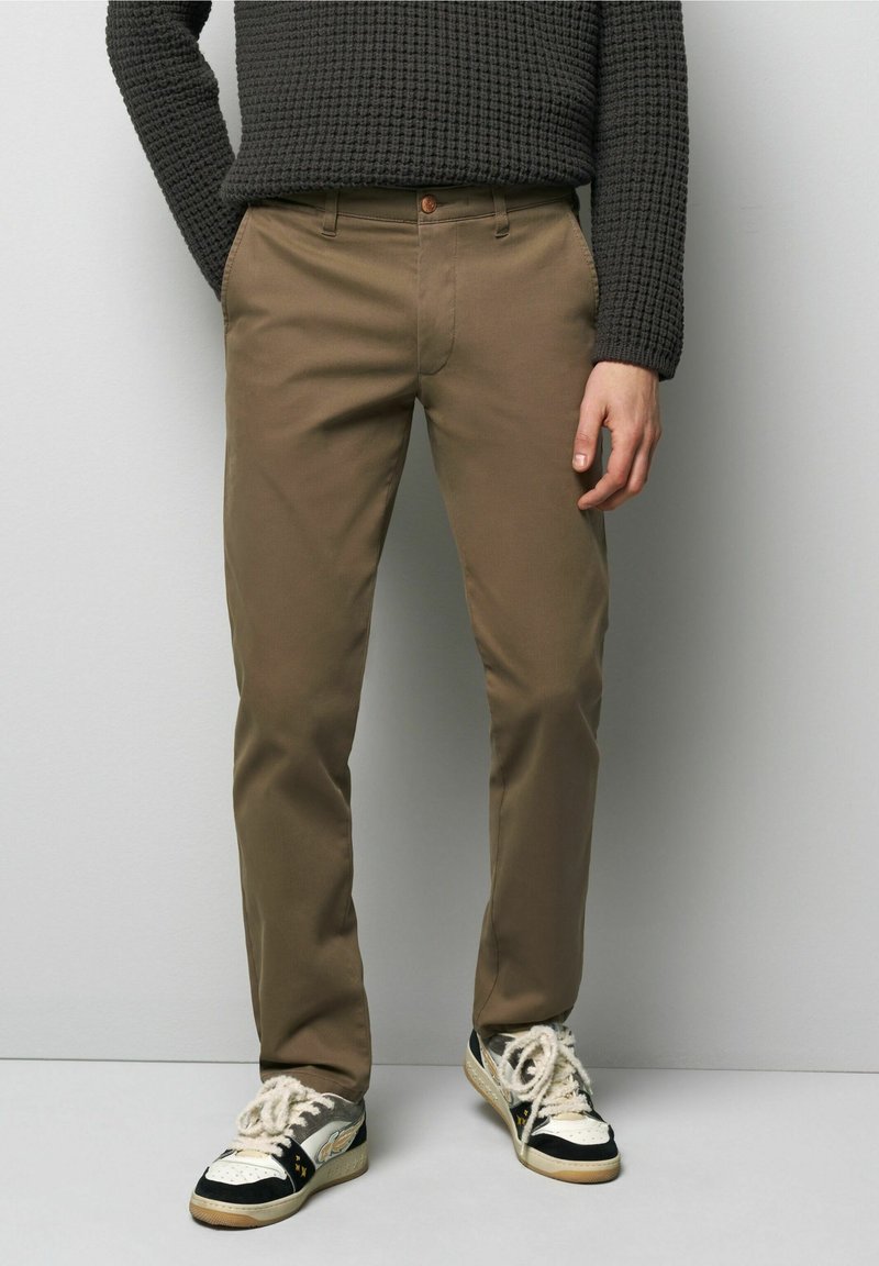 Bruine slim-fit broek van katoen met een gladde textuur, voorzien van zijzakken en een enkele knoopsluiting, gedragen met zwart-witte sneakers.