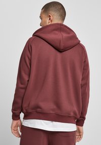 Burgundy huppari, jossa on tilava huppu, ribbaus hihansuissa ja helmassa sekä pehmeä tekstuuri. Ominaisuuksina kerroksellinen ilme, jossa alla valkoinen paita.