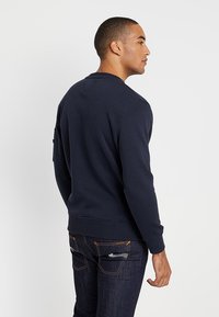 Sudadera azul marino de mezcla de algodón, con cuello redondo, puños acanalados y un detalle de parche en la parte posterior. Combinada con jeans de mezclilla oscura.